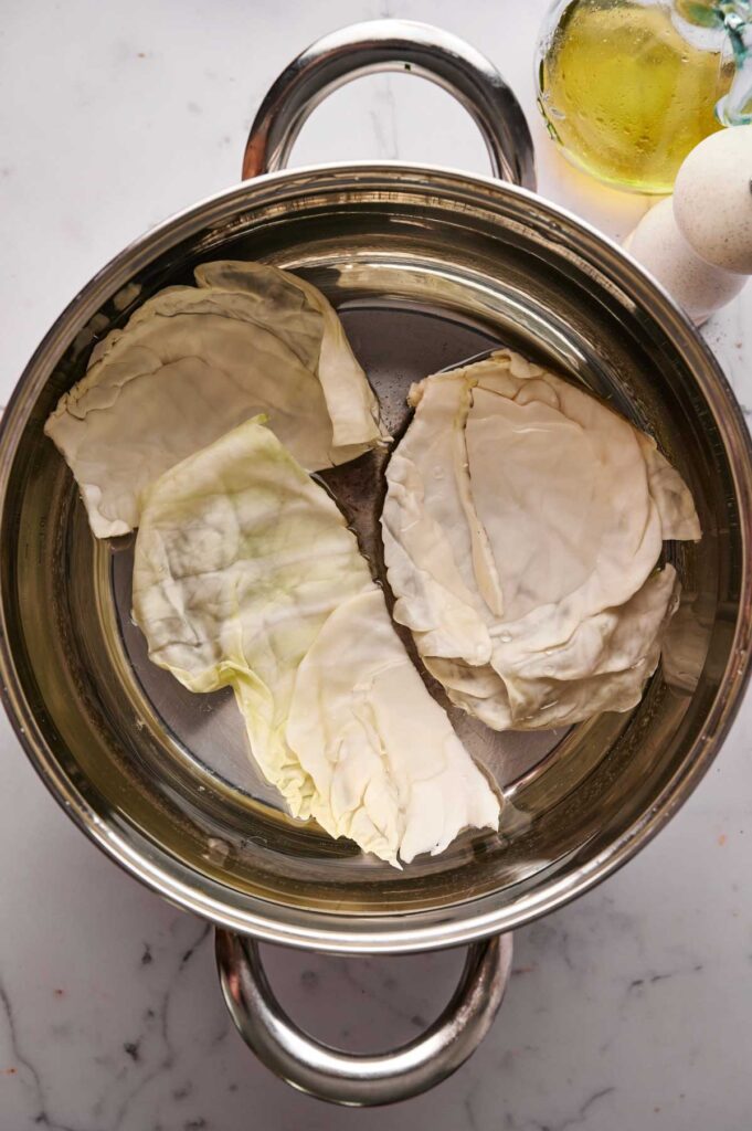 Three cabbage leaves are placed inside a stainless steel pot on a marble countertop, hinting at the start of delicious Stuffed Cabbage Rolls, with a bottle of oil and an egg visible nearby.