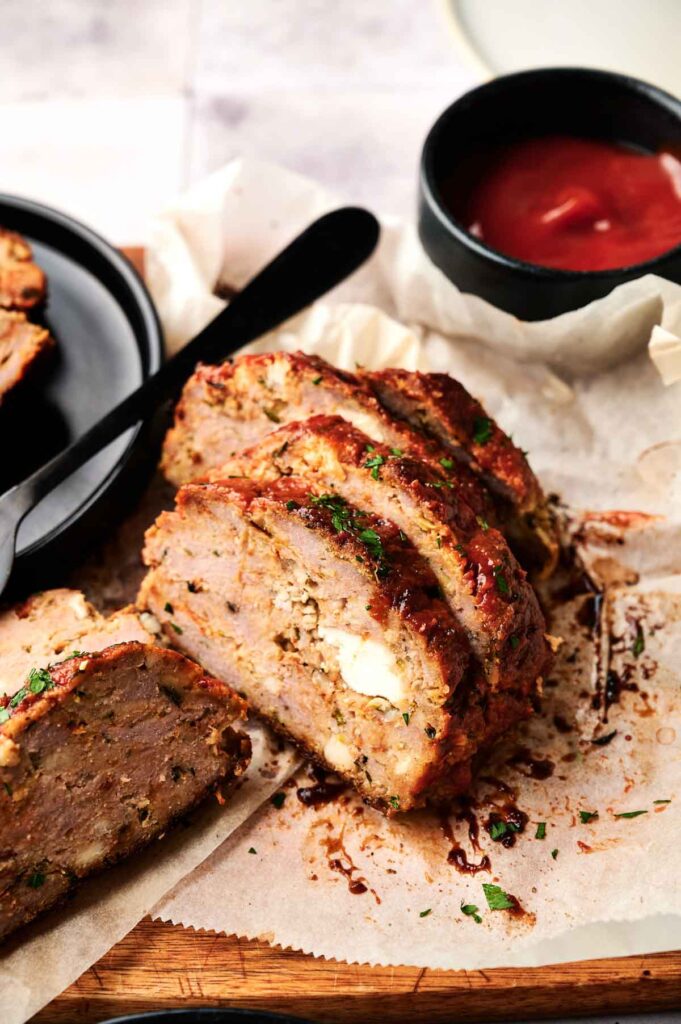 Sliced turkey meatloaf on parchment paper with a bowl of ketchup and a black plate in the background.