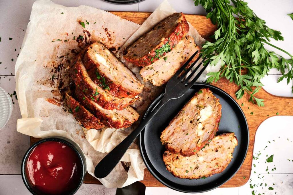 Sliced turkey meatloaf with herbs served on parchment paper, accompanied by a small bowl of ketchup and fresh parsley on a wooden cutting board.