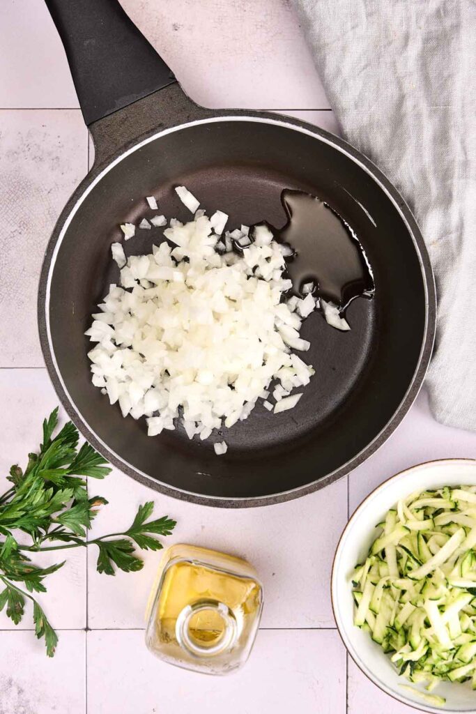 A frying pan with chopped onions and oil, surrounded by fresh parsley, a bowl of shredded zucchini for turkey meatloaf, and a small bottle of oil on a tiled surface.
