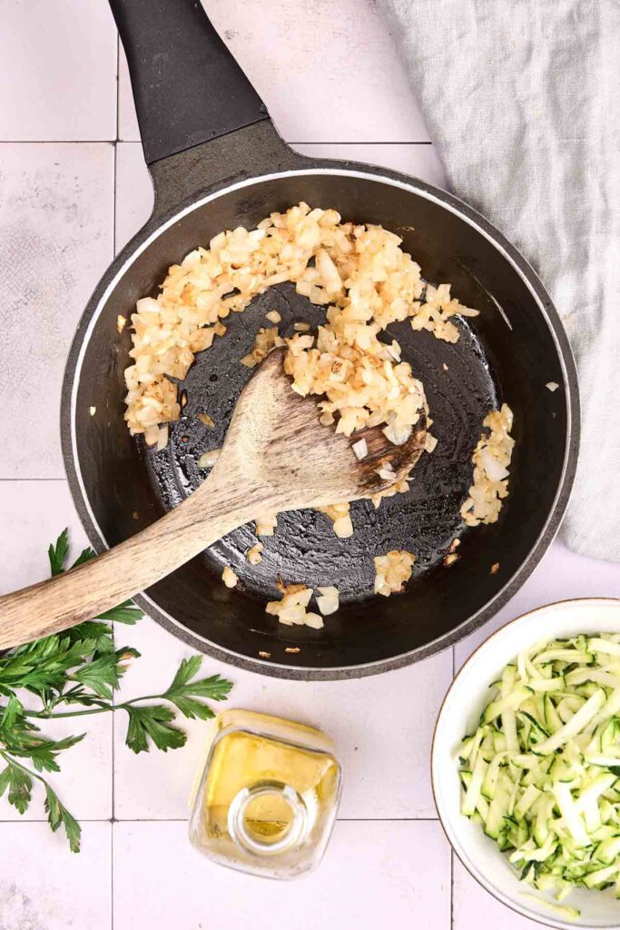 Chopped onions saut&eacute;ing in a black skillet with a wooden spoon, next to fresh parsley, a bowl of shredded vegetables, and a bottle of olive oil&mdash;perfect for preparing Turkey Meatloaf on a tiled surface.