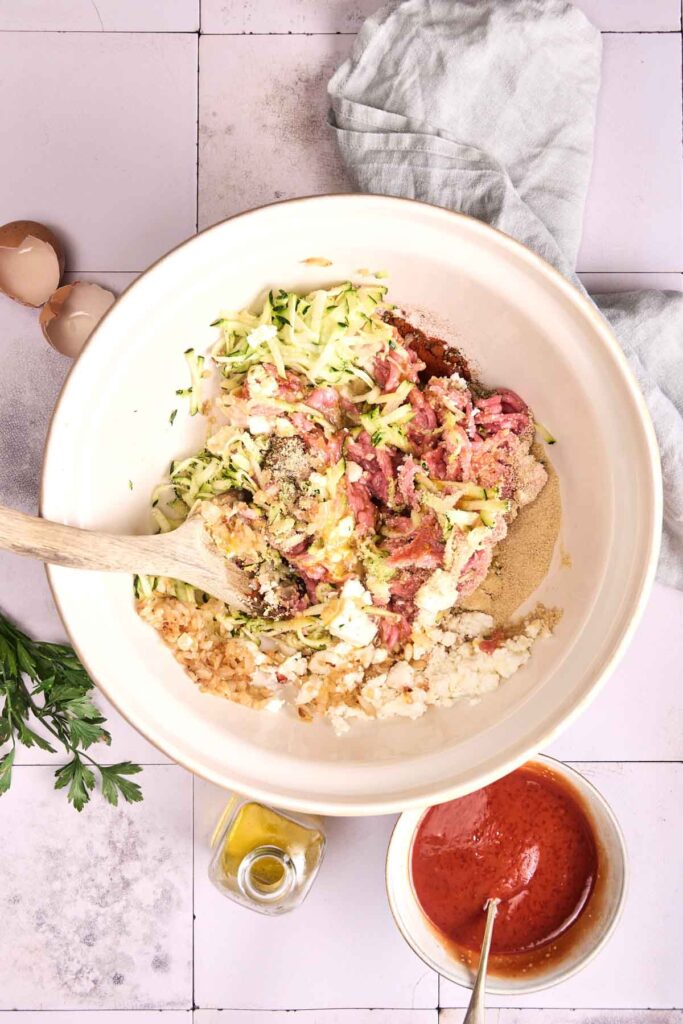 Large bowl contains Turkey Meatloaf mix&mdash;ground meat, shredded zucchini, onions, breadcrumbs, egg, and seasonings&mdash;with a wooden spoon. Nearby are cracked eggs, parsley, olive oil, and a bowl of tomato sauce.