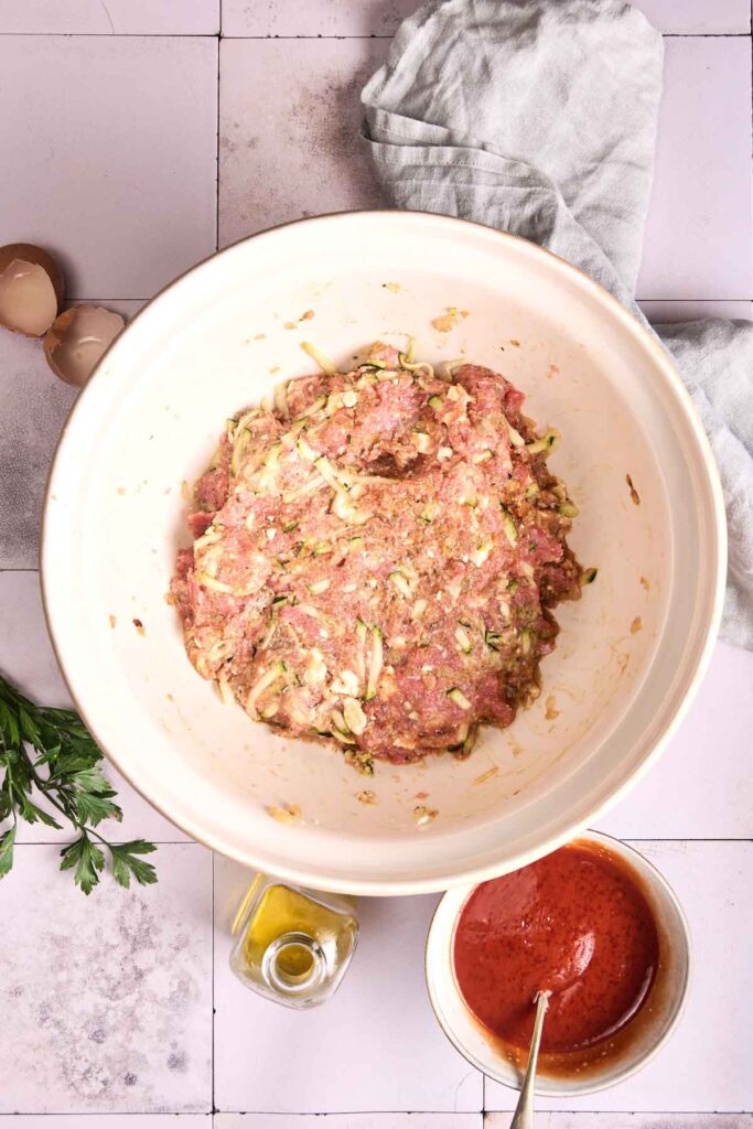 Large mixing bowl with Turkey Meatloaf mixture and seasonings, surrounded by cracked eggs, tomato sauce in a small bowl, olive oil, fresh parsley, and a cloth napkin.
