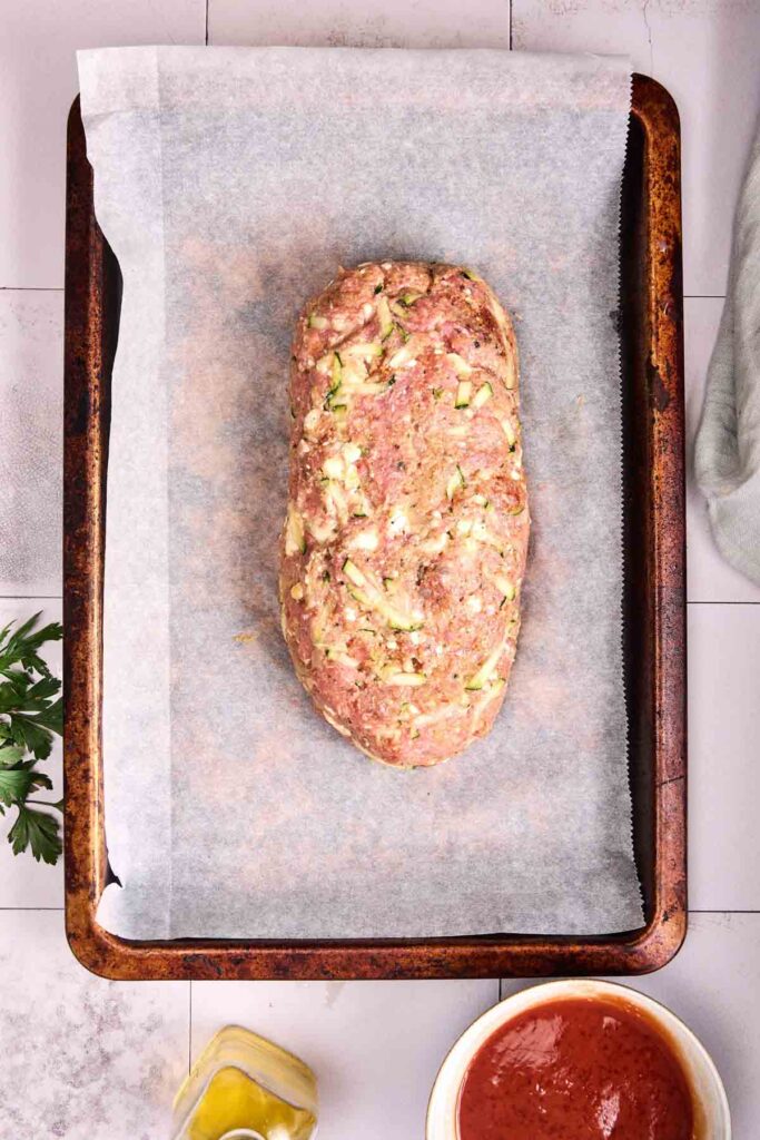 Uncooked turkey meatloaf, shaped into a loaf, sits on parchment paper on a baking tray, with bowls of sauce and oil nearby.