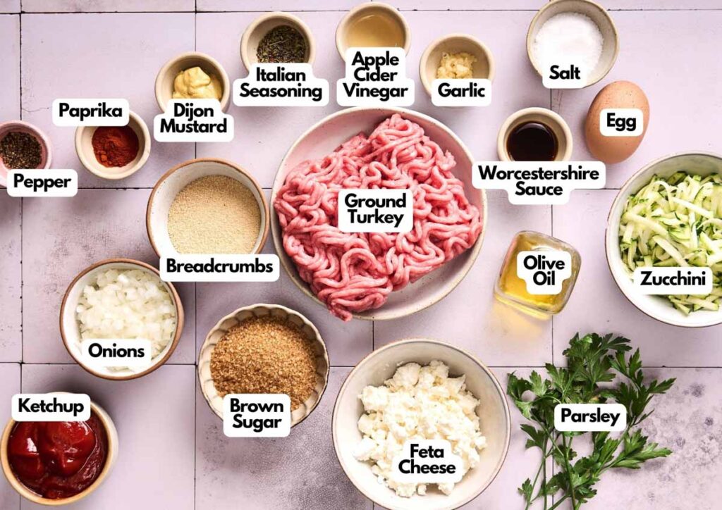 A variety of Turkey Meatloaf ingredients&mdash;ground turkey, eggs, breadcrumbs, onions, zucchini, feta cheese, seasonings, and sauces&mdash;are neatly arranged on a kitchen counter.