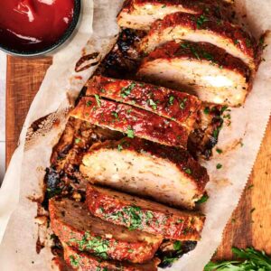 Sliced turkey meatloaf topped with herbs on parchment paper, served on a wooden board with a small bowl of ketchup nearby.