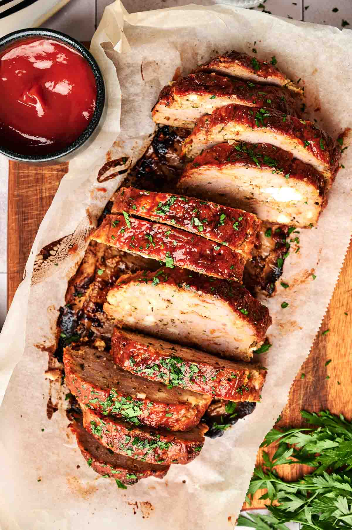 Sliced turkey meatloaf topped with herbs on parchment paper, served on a wooden board with a small bowl of ketchup nearby.