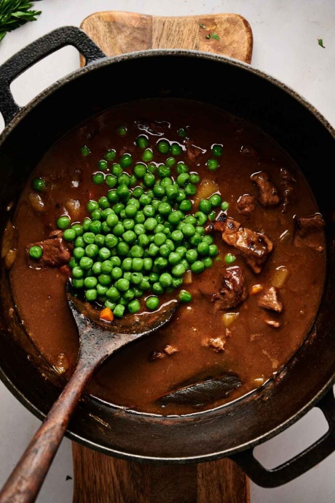 A pot of Vegetable Beef Soup with chunks of meat, vegetables, and green peas is being stirred with a wooden spoon.