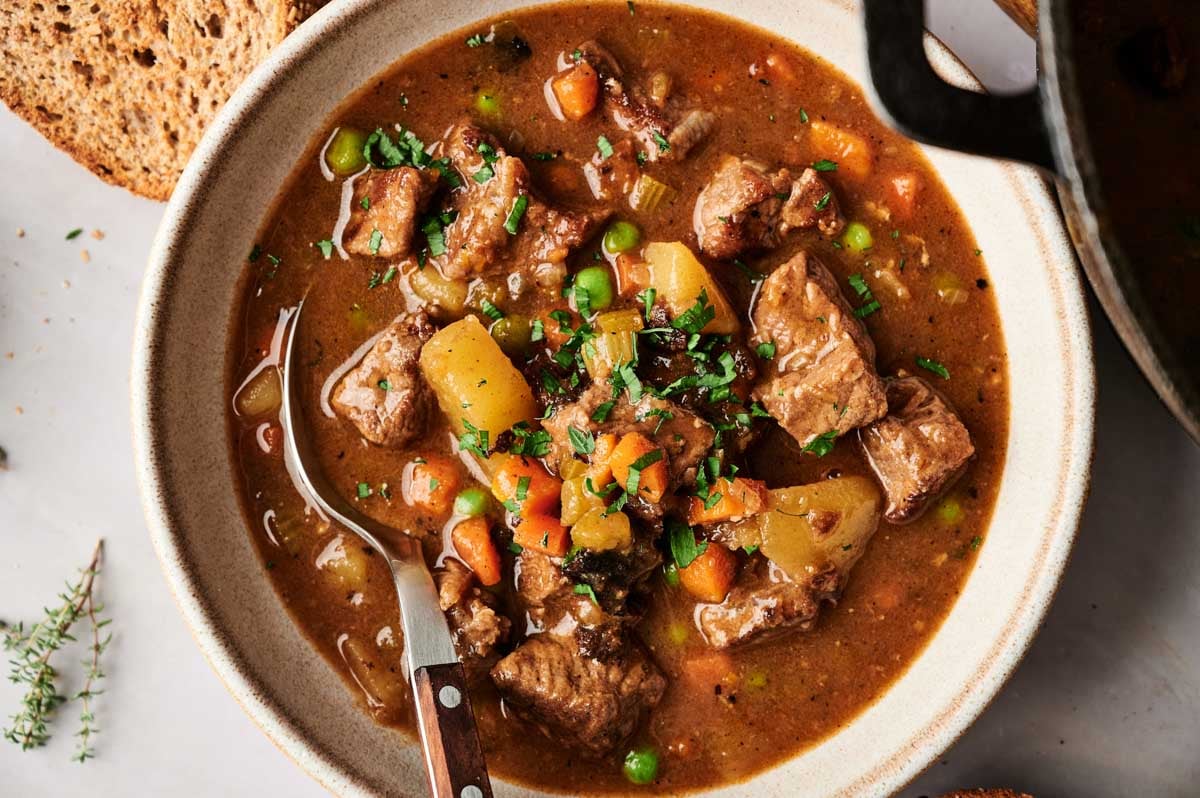 A bowl of hearty Vegetable Beef Soup, filled with chunks of beef, potatoes, carrots, peas, and herbs in a thick broth, served alongside a slice of bread and a fork.