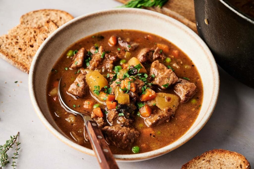 A bowl of hearty Vegetable Beef Soup, filled with chunks of beef, potatoes, carrots, and peas in a rich broth, served with slices of bread on the side.