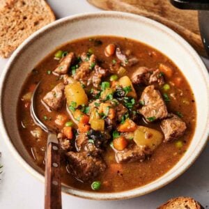 A bowl of hearty Vegetable Beef Soup with chunks of beef, potatoes, carrots, and peas in a savory broth, served with slices of bread on the side.