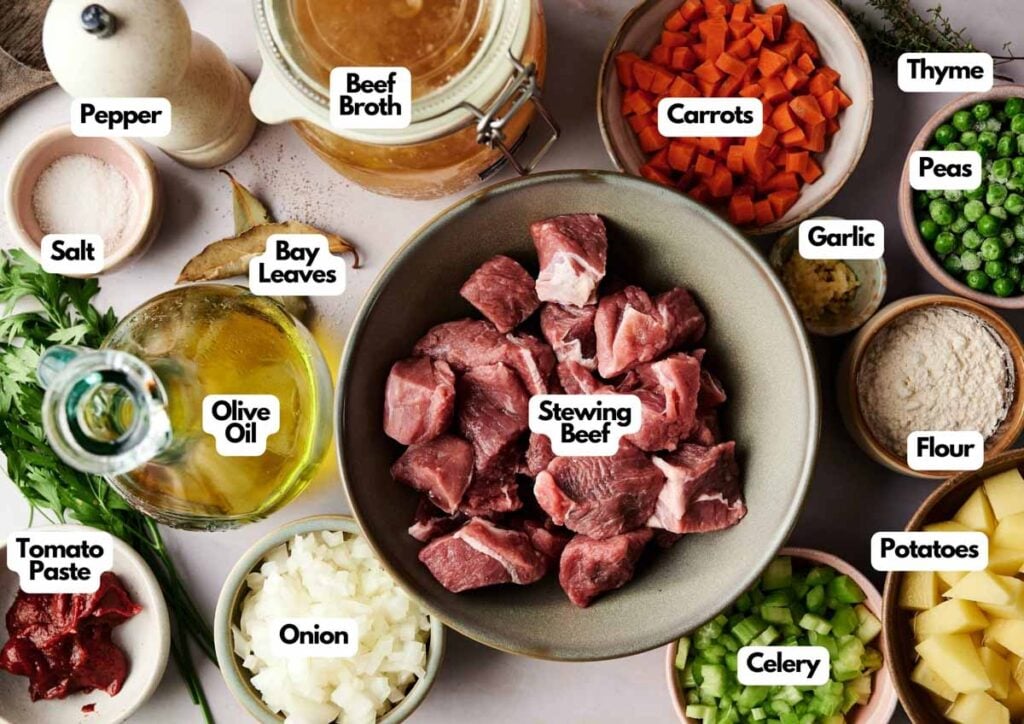 A bowl of raw stewing beef is surrounded by labeled ingredients for Vegetable Beef Soup: olive oil, tomato paste, onion, beef broth, bay leaves, salt, pepper, carrots, thyme, peas, garlic, flour, potatoes, and celery.