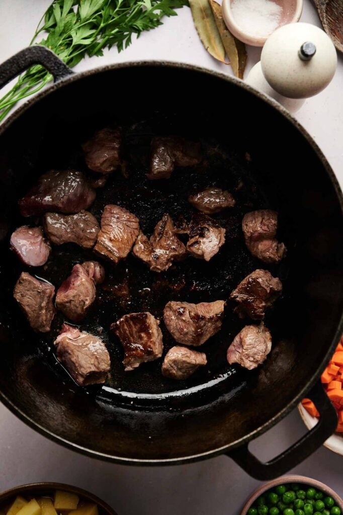 Chunks of beef brown in a black cast iron pot, surrounded by carrots, peas, and herbs&mdash;classic ingredients for a hearty Vegetable Beef Soup&mdash;on a light countertop.