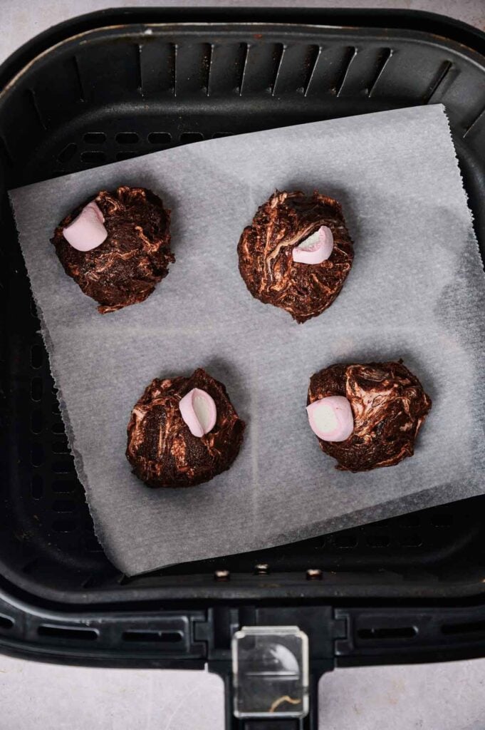 Four unbaked Air Fryer Marshmallow Swirl Cookies dough balls with marshmallow pieces on top are arranged on parchment paper inside an air fryer basket.