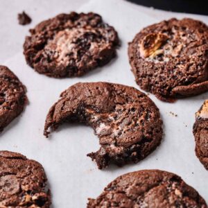 Six Air Fryer Marshmallow Swirl Cookies, one with a bite taken out, rest on a sheet of parchment paper with a dark container partially visible in the background.