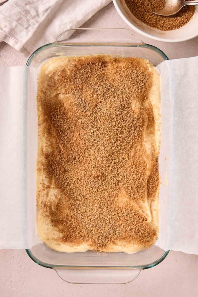 A glass baking dish lined with parchment paper holds unbaked Amish Cinnamon Cake dough topped with a generous layer of brown cinnamon sugar. Nearby, a bowl of cinnamon sugar and a spoon are ready for the next step.