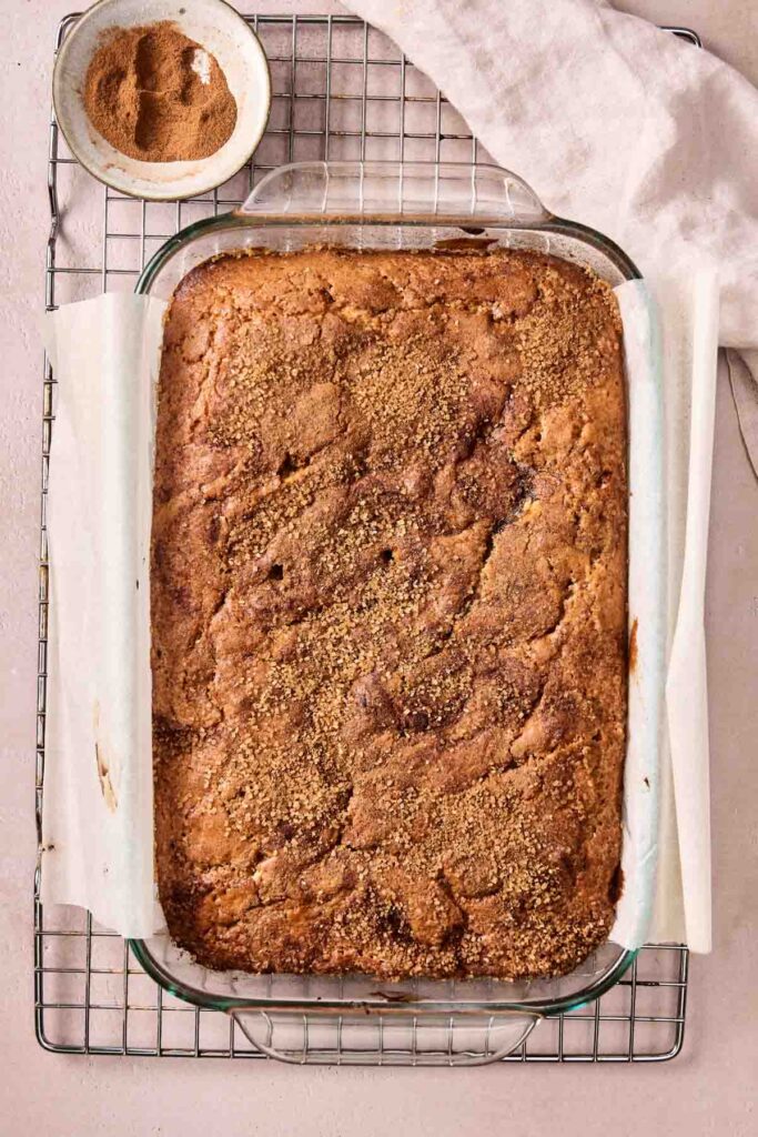A rectangular glass baking dish containing a baked Amish Cinnamon Cake with a crumbly cinnamon topping sits on a cooling rack, with a small bowl of cinnamon sugar nearby.