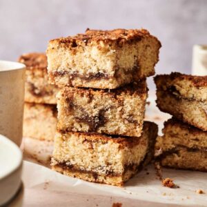 A stack of three square Amish Cinnamon Cake bars with a cinnamon layer, surrounded by more bars and white ceramic cups on a light surface.