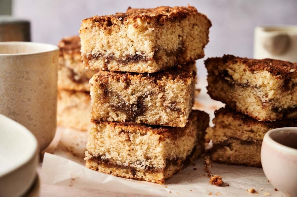 Three pieces of crumbly Amish Cinnamon Cake with a cinnamon swirl are stacked on parchment paper, with more cake pieces and cups in the background.