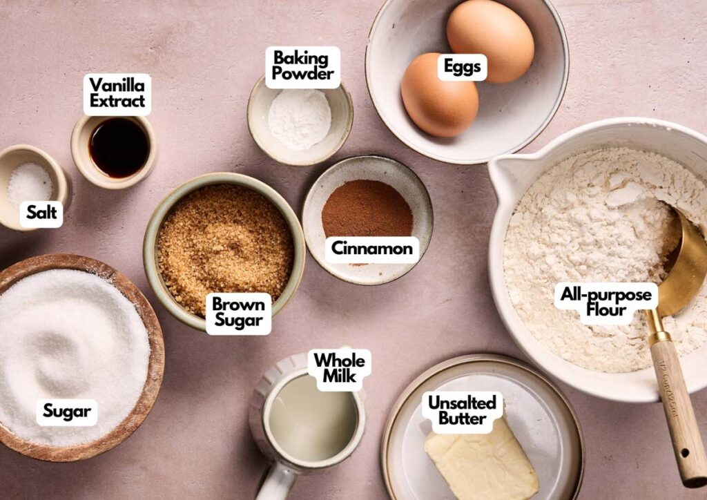 Various labeled baking ingredients for Amish Cinnamon Cake, including eggs, flour, butter, milk, sugars, cinnamon, baking powder, salt, and vanilla extract, are arranged on a light pink surface.
