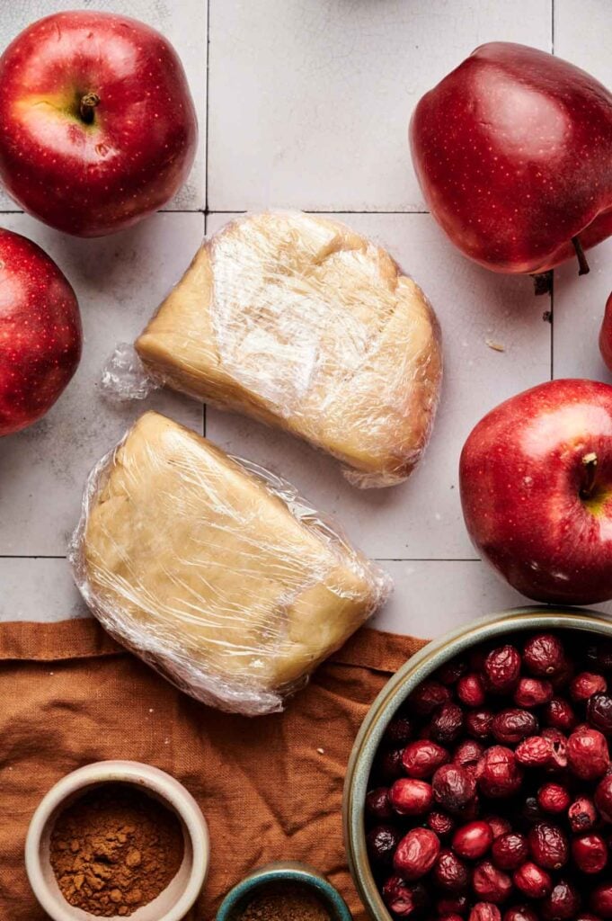 Wrapped dough, whole red apples, a bowl of fresh cranberries, and small bowls of cinnamon and brown sugar are arranged on a tiled surface&mdash;perfect ingredients for crafting a homemade Apple Cranberry Pie.