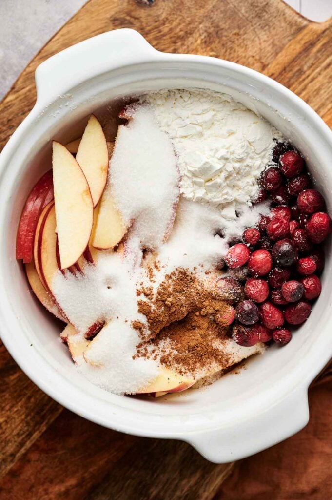White bowl containing sliced apples, cranberries, flour, sugar, and cinnamon&mdash;perfect ingredients for Apple Cranberry Pie&mdash;placed on a wooden surface.