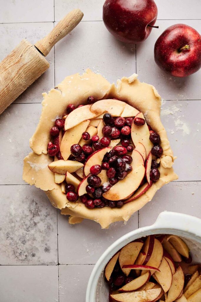 Unbaked Apple Cranberry Pie with sliced apples and cranberries in a pastry crust, surrounded by a rolling pin, two red apples, and a mixing bowl on a tiled surface.