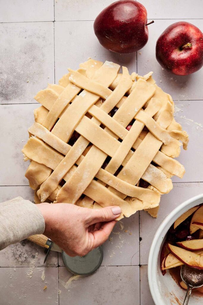 A hand crimps the edge of an unbaked lattice Apple Cranberry Pie on a tiled countertop, with two apples and a bowl of sliced apples nearby.