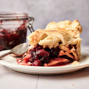 A slice of Apple Cranberry Pie with a flaky crust is on a plate, with a jar of fruit filling and a spoon in the background.
