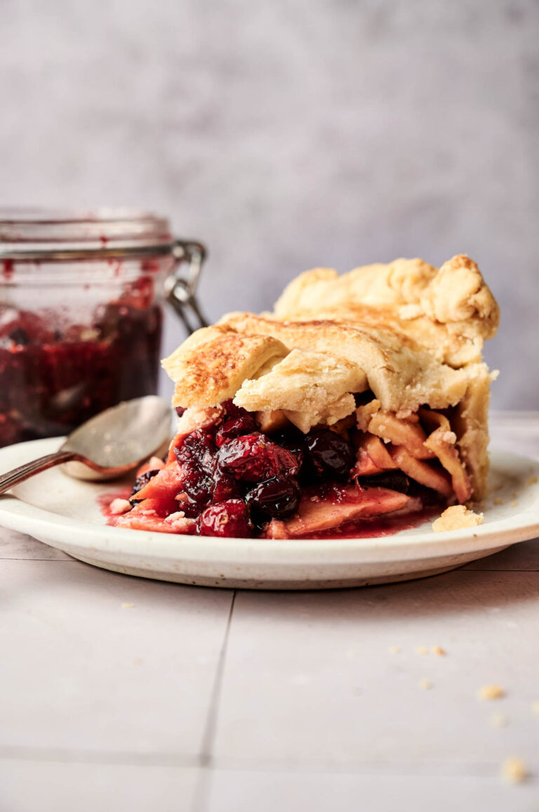 A slice of Apple Cranberry Pie with a flaky crust is on a plate, with a jar of fruit filling and a spoon in the background.