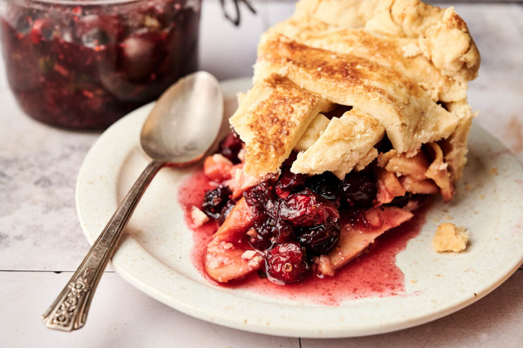 A slice of Apple Cranberry Pie with a golden crust sits on a white plate next to a spoon, its tangy berry and apple filling visible, with a jar of extra pie filling in the background.