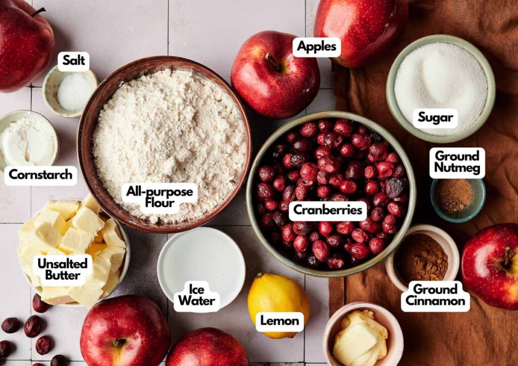 Assorted baking ingredients for Apple Cranberry Pie, including apples, cranberries, flour, butter, ice water, lemon, salt, cornstarch, sugar, nutmeg, and cinnamon arranged on a countertop.