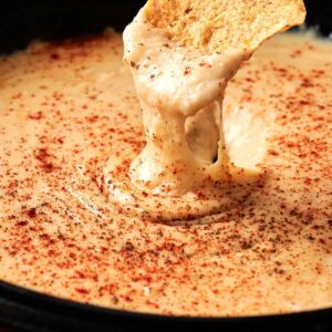 A hand dips a tortilla chip into a skillet of melted Beer Cheese Dip, topped with paprika.
