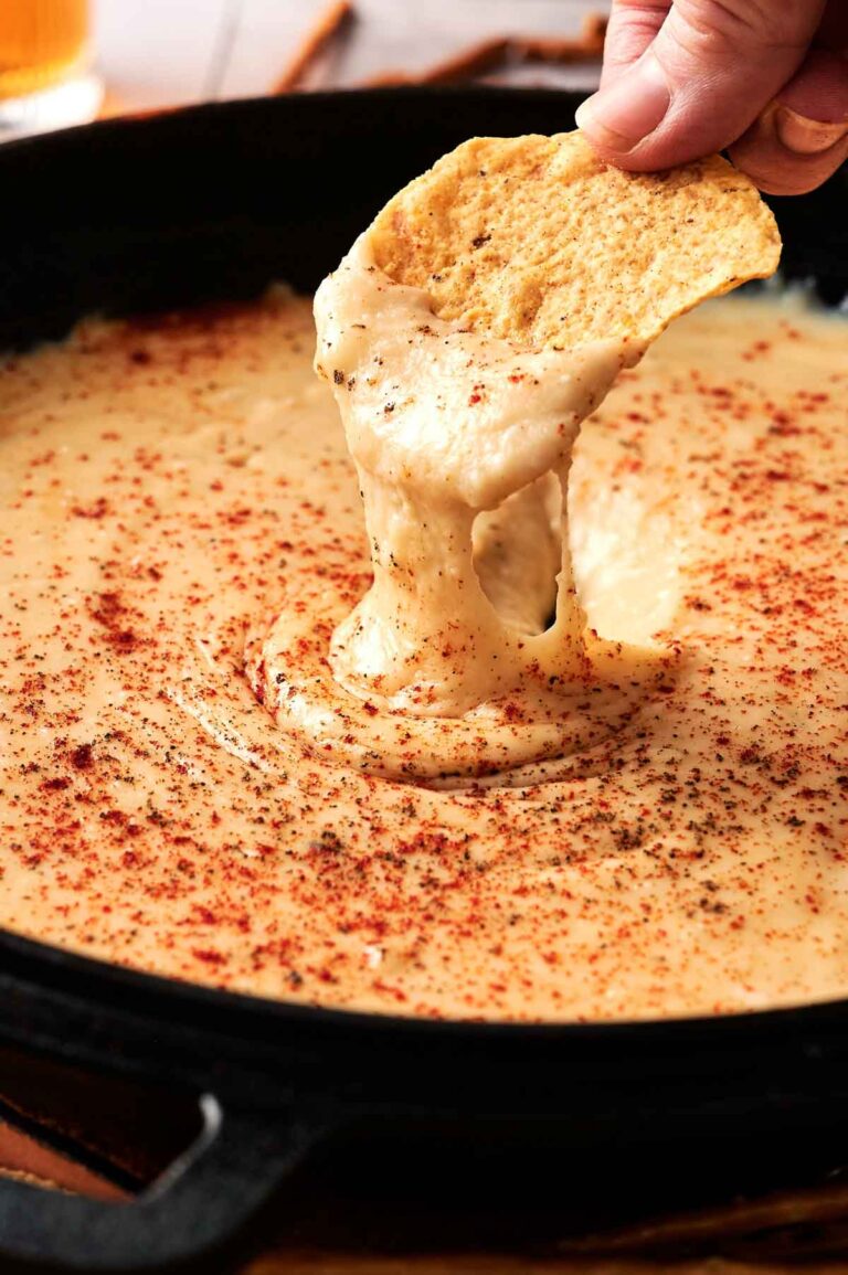 A hand dips a tortilla chip into a skillet of melted Beer Cheese Dip, topped with paprika.