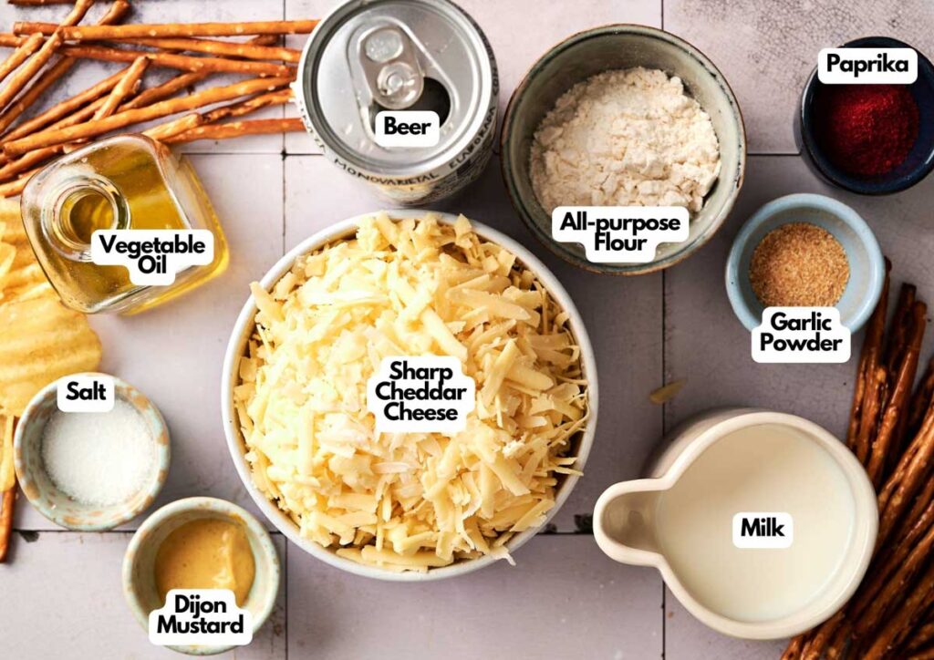 Top-down view of labeled ingredients for Beer Cheese Dip, featuring sharp cheddar cheese, beer, flour, milk, vegetable oil, Dijon mustard, salt, paprika, and garlic powder.
