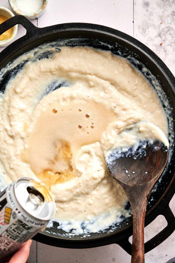 A can of beer is being poured into a creamy mixture in a black skillet to make delicious Beer Cheese Dip, with a wooden spoon stirring the contents.