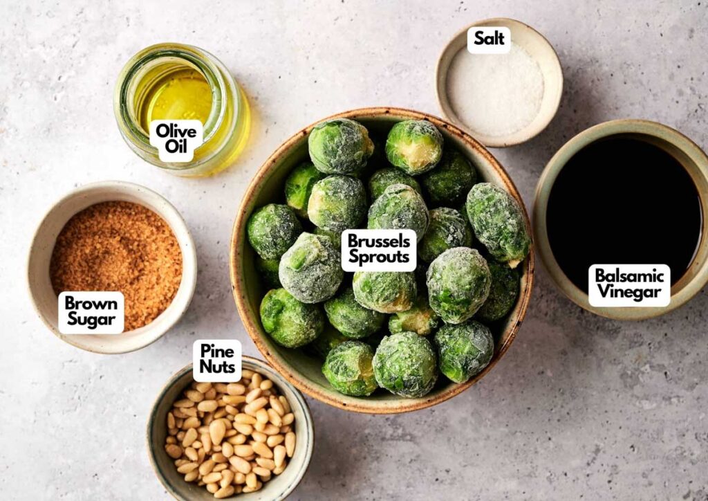 A bowl of frozen Brussels sprouts surrounded by bowls of olive oil, salt, balsamic vinegar, brown sugar, and pine nuts&mdash;perfect ingredients for Brussels Sprouts with Balsamic Glaze&mdash;sit on a light surface.