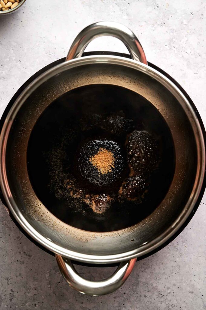 A stainless steel pot containing a dark liquid for Brussels Sprouts with Balsamic Glaze, with granulated brown sugar in the center, sits on a light gray countertop.