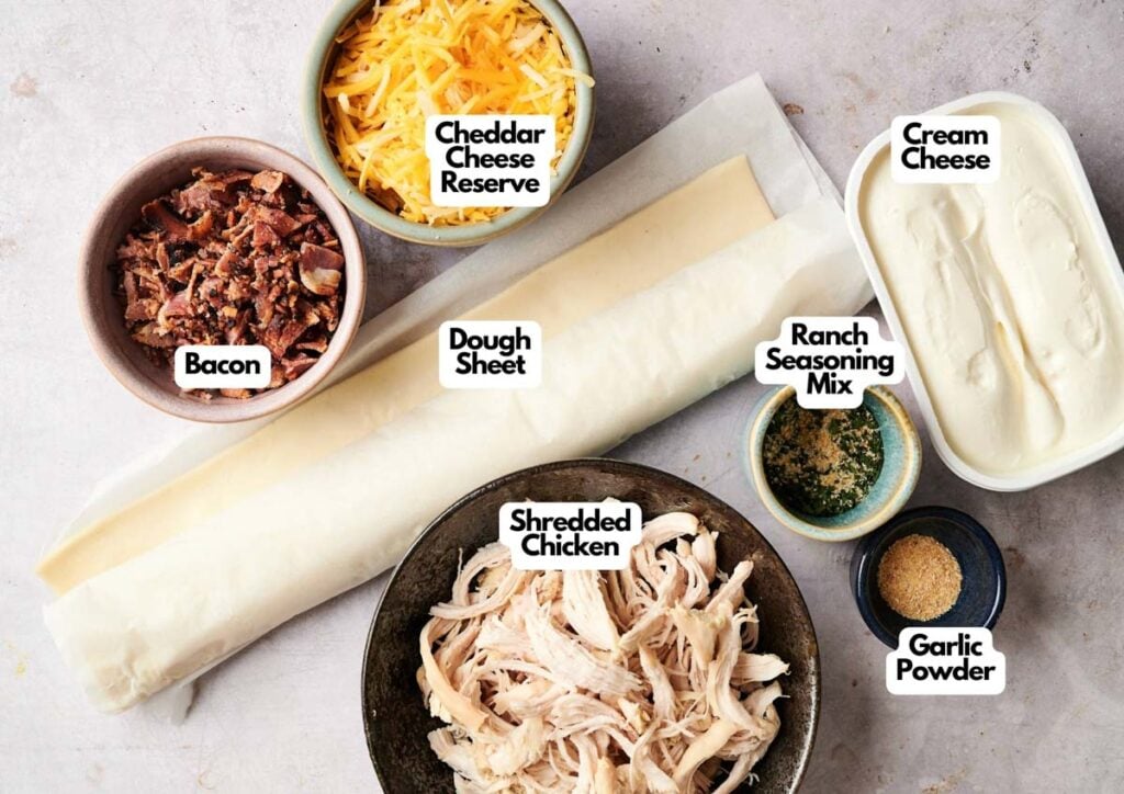 Ingredients for Chicken Pinwheels are laid out on a countertop: dough sheet, shredded chicken, bacon, cheddar cheese, cream cheese, ranch seasoning mix, and garlic powder.