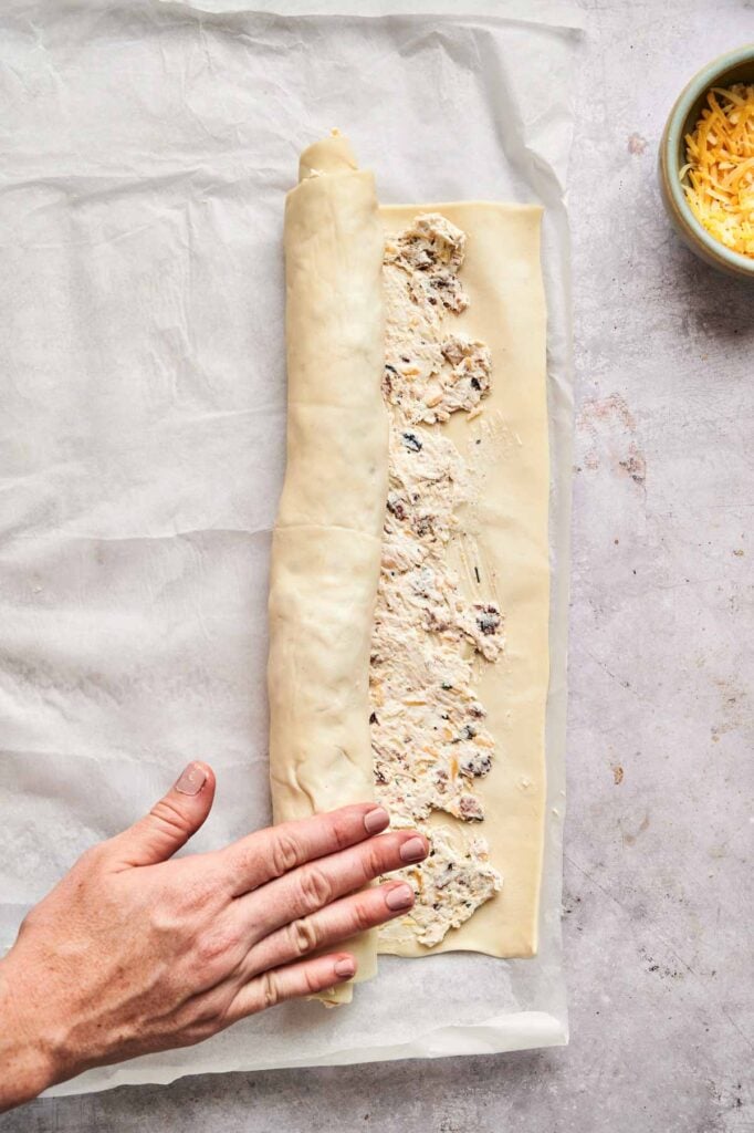 A hand rolls up puff pastry filled with a creamy mixture to make delicious Chicken Pinwheels on parchment paper; a bowl of grated cheese sits nearby.