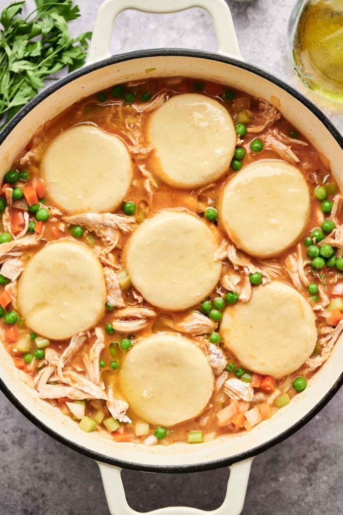 A pot of Chicken and Biscuits stew, filled with tender chicken and vegetables, is topped with six round biscuit dough pieces, ready to be baked.