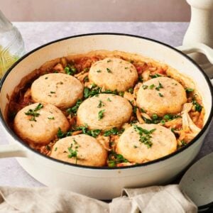 A white pot filled with chicken and vegetable stew topped with six round dumplings, reminiscent of classic chicken and biscuits, garnished with chopped parsley.