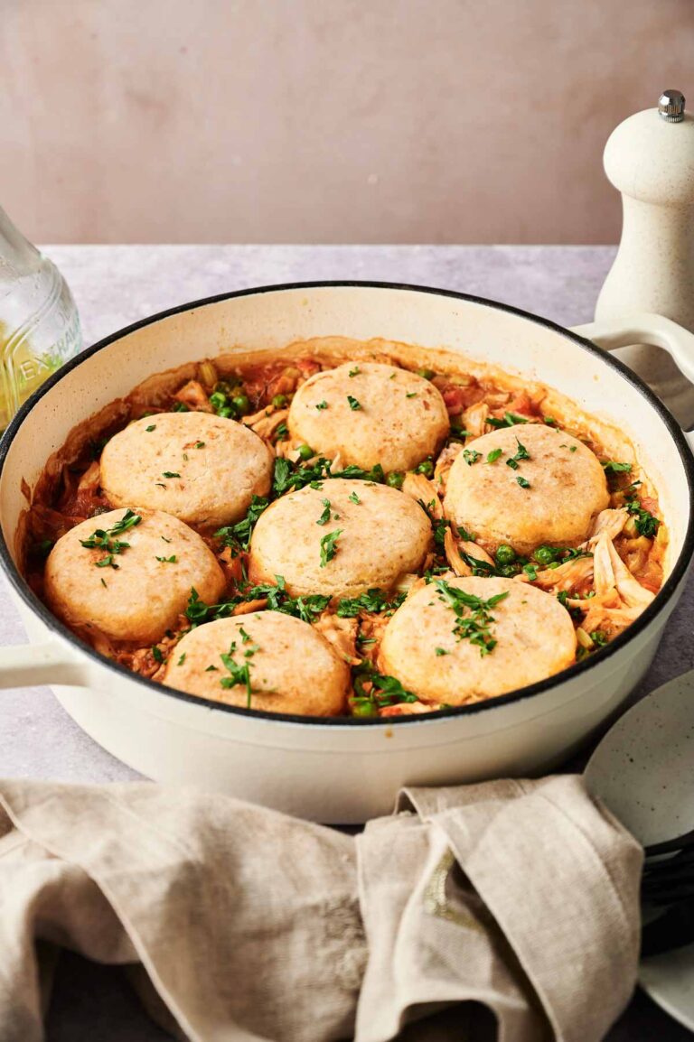 A white pot filled with chicken and vegetable stew topped with six round dumplings, reminiscent of classic chicken and biscuits, garnished with chopped parsley.