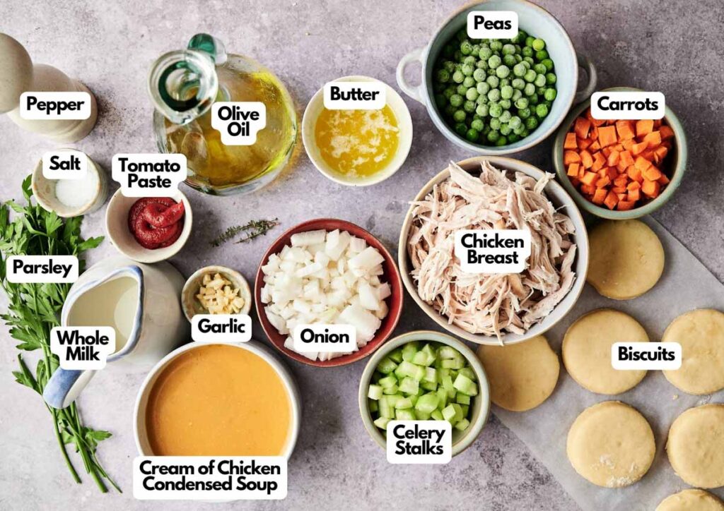 Ingredients for Chicken and Biscuits pot pie arranged on a surface, including chicken breast, biscuits, peas, carrots, celery, onion, garlic, butter, olive oil, cream of chicken soup, milk, and seasonings.