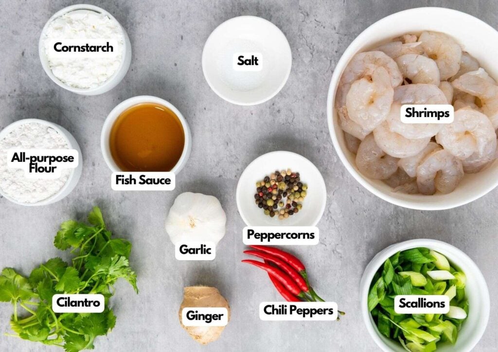Top-down view of labeled ingredients for Chinese Salt and Pepper Shrimp on a gray surface, including shrimps, salt, cornstarch, flour, fish sauce, garlic, peppercorns, chili peppers, scallions, ginger, and cilantro.