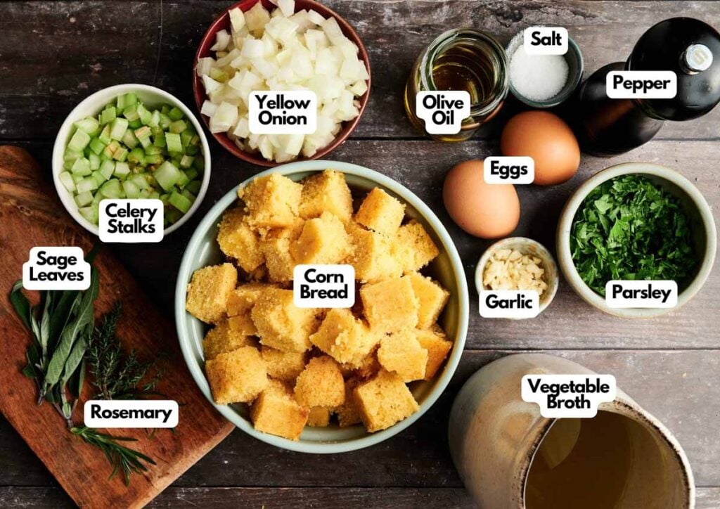 Overhead view of ingredients for Cornbread Stuffing, featuring cubed cornbread, eggs, chopped vegetables, fresh herbs, garlic, olive oil, salt, pepper, and vegetable broth.