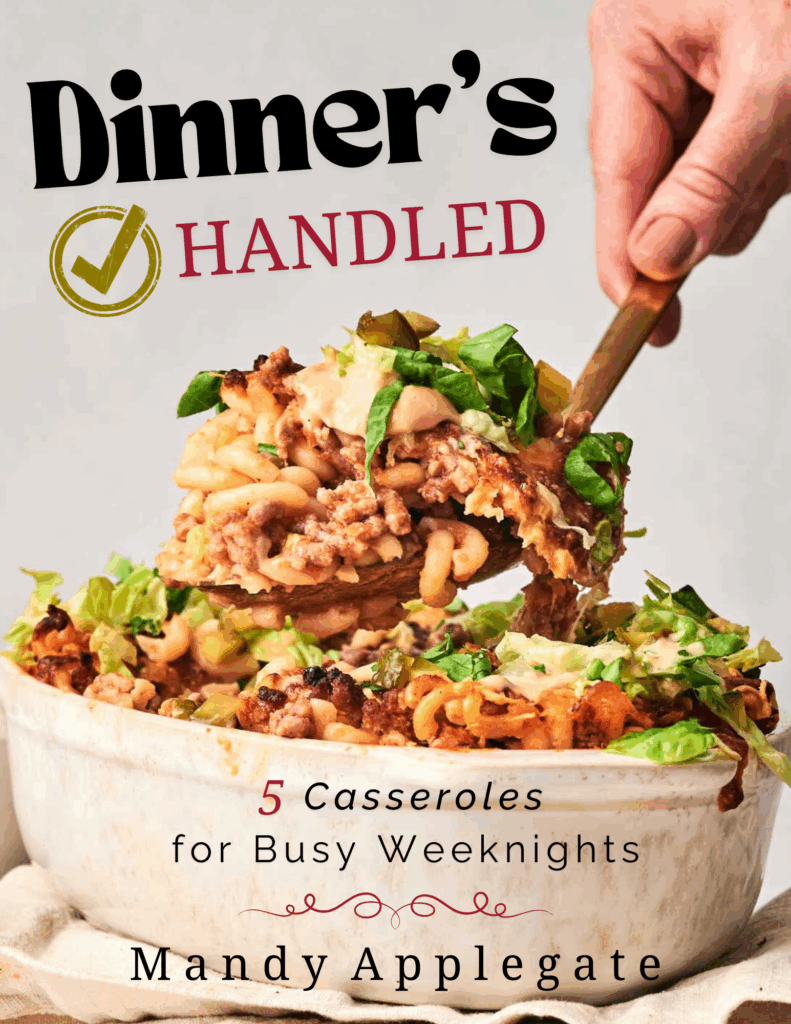 A hand serves a portion of casserole from a dish filled with baked pasta and vegetables; text on the image promotes a cookbook titled “Dinner’s Handled” by Mandy Applegate.