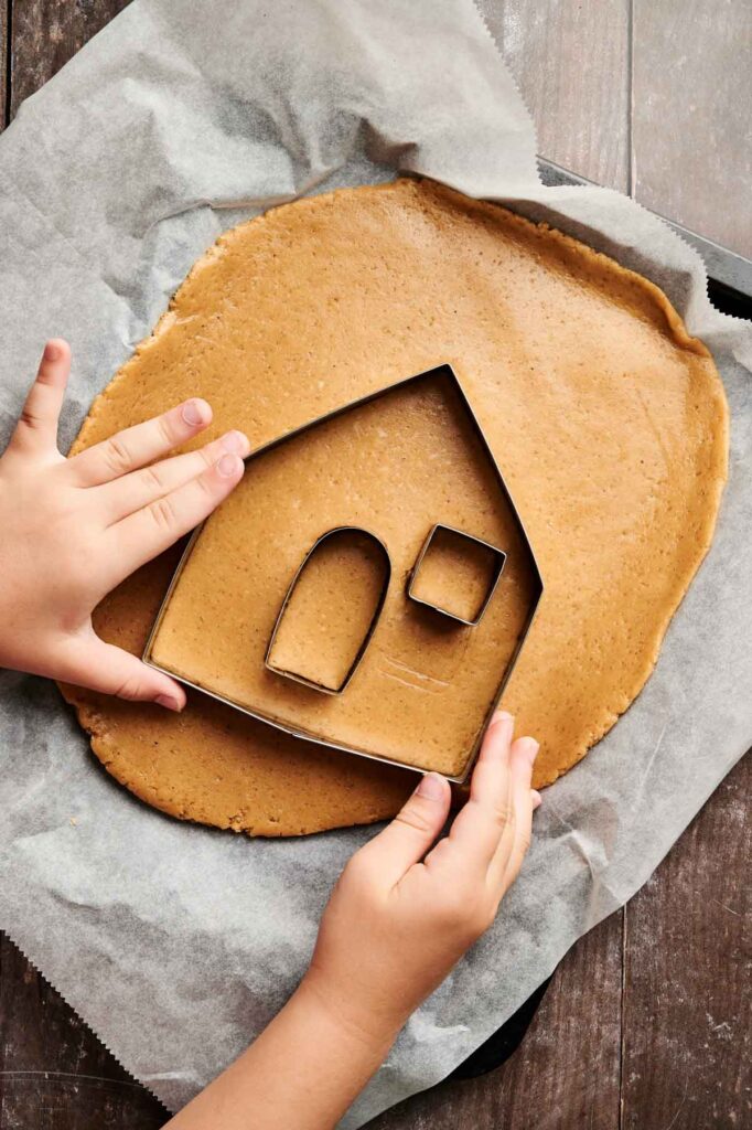 A child's hands press a house-shaped cookie cutter into rolled gingerbread dough, ready to create the start of a festive Gingerbread House on parchment paper.