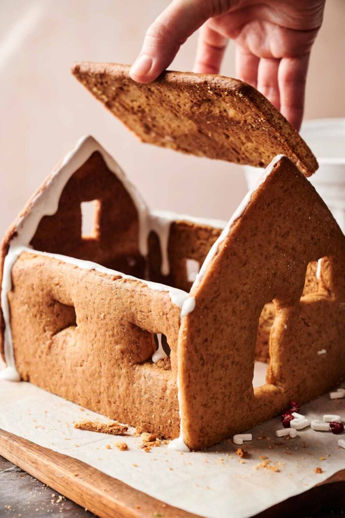 A hand lifts the roof piece onto a partially assembled Gingerbread House, with white icing neatly outlining the edges.