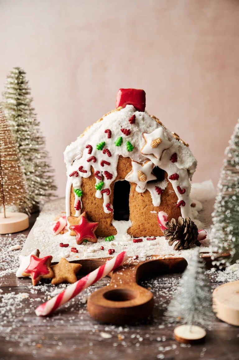 A charming Gingerbread House decorated with white icing, star-shaped cookies, candy canes, and artificial trees sits on a snowy surface.